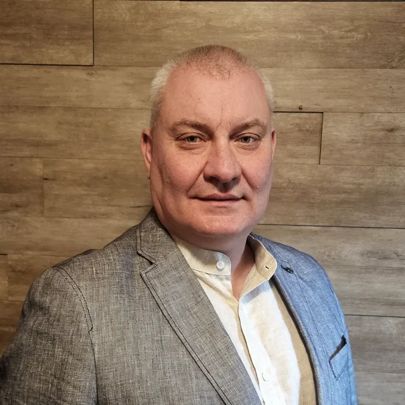 Porträtfoto eines Mannes mittleren Alters mit kurzen grauen Haaren und leichtem Lächeln. Er trägt ein weißes Hemd mit offenem Kragen und ein grau meliertes Sakko. Der Hintergrund besteht aus einer Wand mit horizontaler Holzpaneelverkleidung in hellem Eicheton. Quadratisches Format, Brustbild.